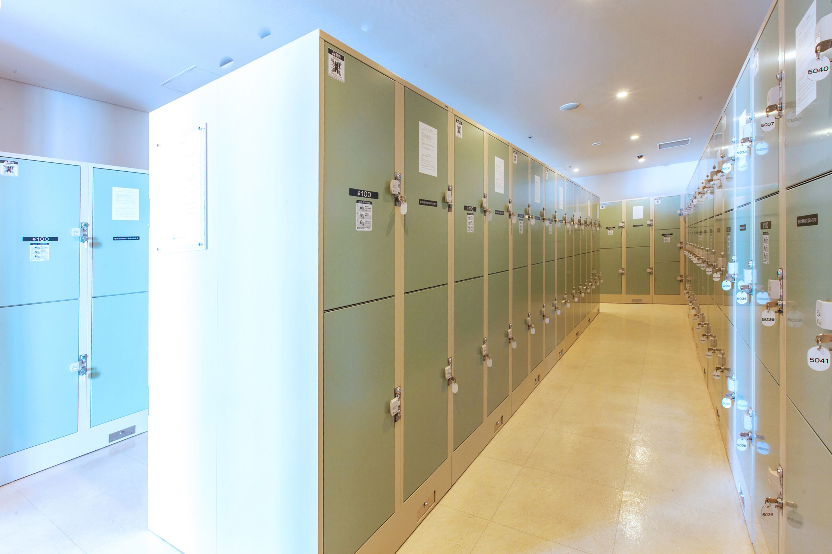 A row of lockers with numbers on doors, a tiled floor, and a ceiling with recessed lighting.