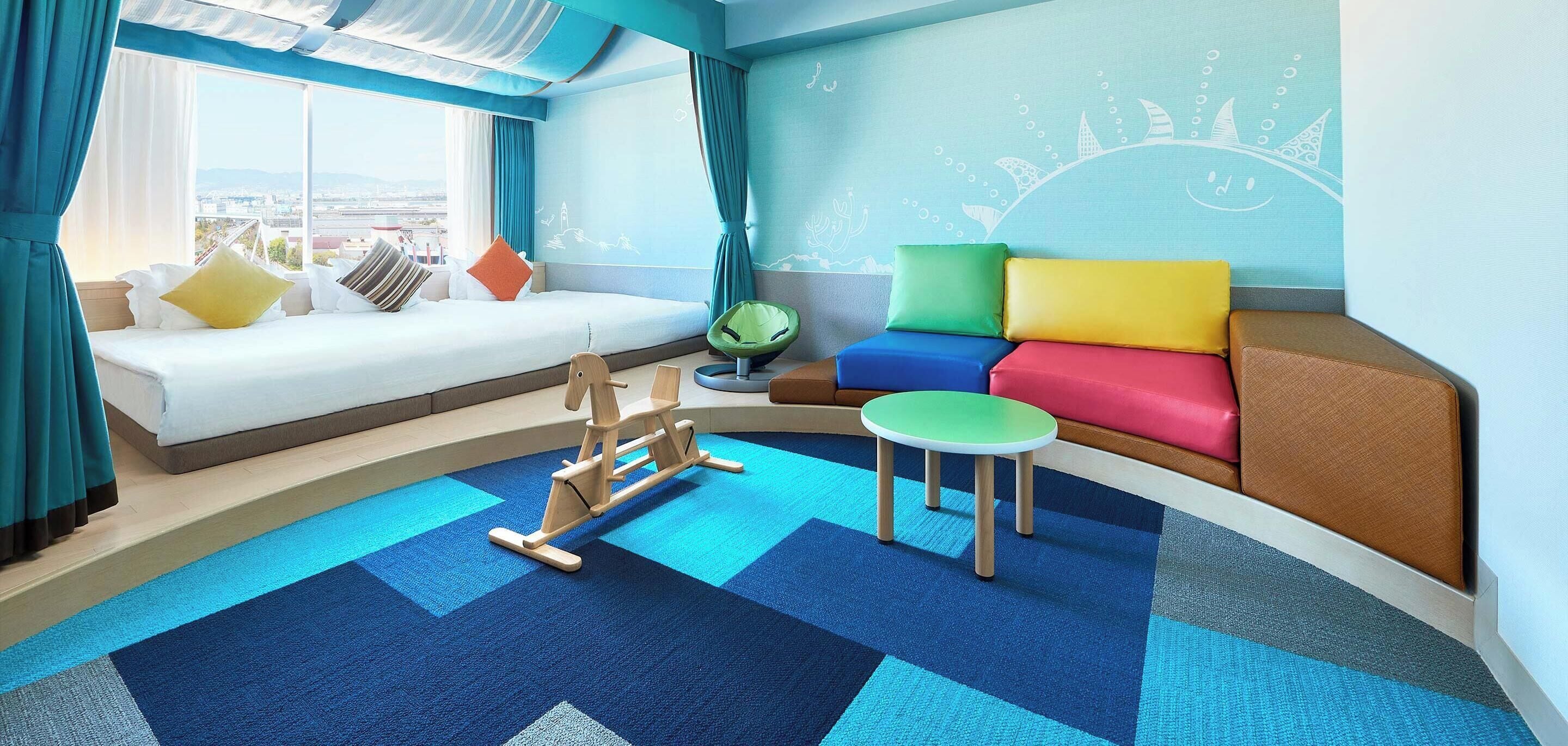 A hotel room with a large window, a sofa with colorful cushions, a small round table, and a rocking horse.