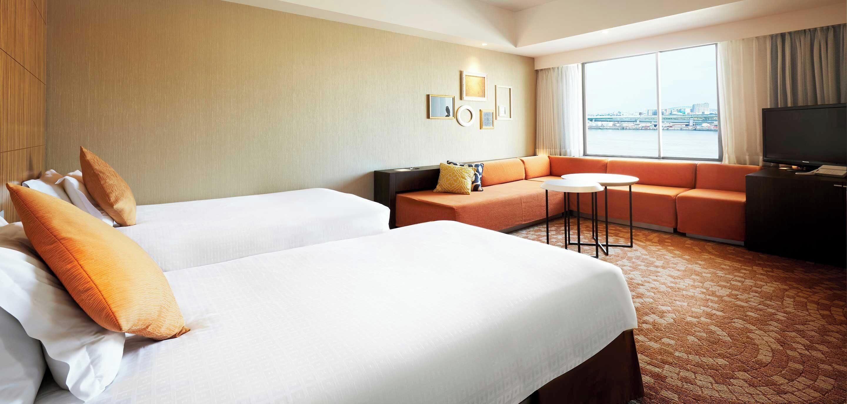Hotelkamer met twee bedden, een bank, een klein tafeltje, een televisie en een groot raam met uitzicht op de stad.