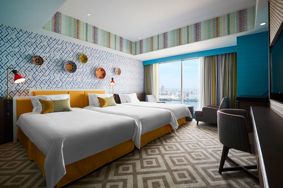 Habitación de hotel con dos camas, un escritorio y un amplio ventanal con vistas a la ciudad.