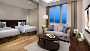 Una habitación de hotel moderna con dos camas, un sofá, una mesa de centro con flores y un ventanal con vista a la ciudad.
