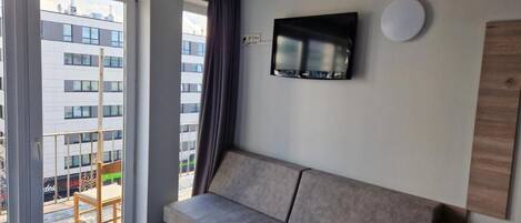 Ein modernes Hotelzimmer mit großem Fenster, einer Couch, einem Flachbildfernseher und einem Holznachttisch.