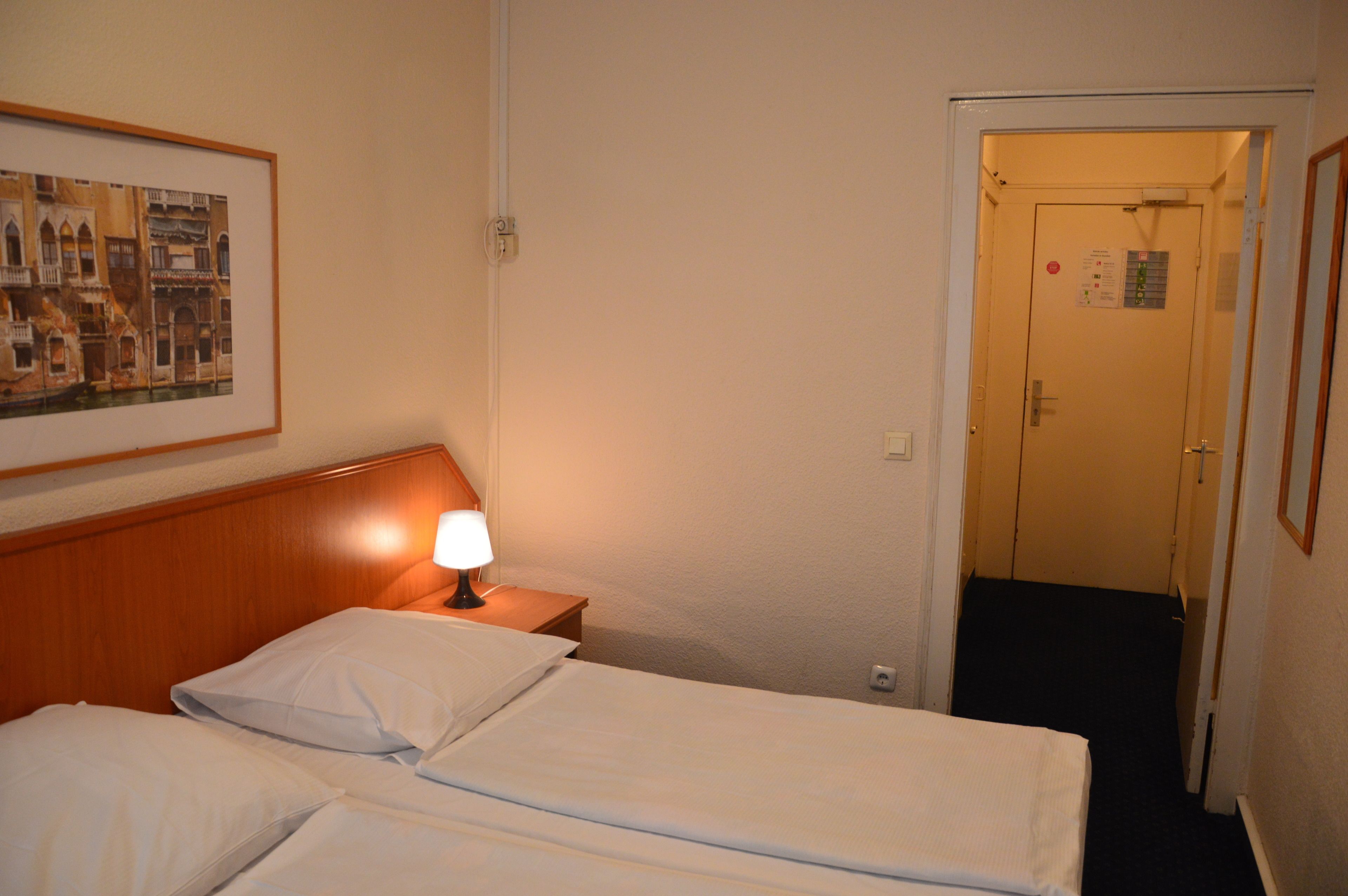 Una habitación de hotel con cama, una mesita de noche con lámpara, un cuadro en la pared y una puerta que da a otra habitación.