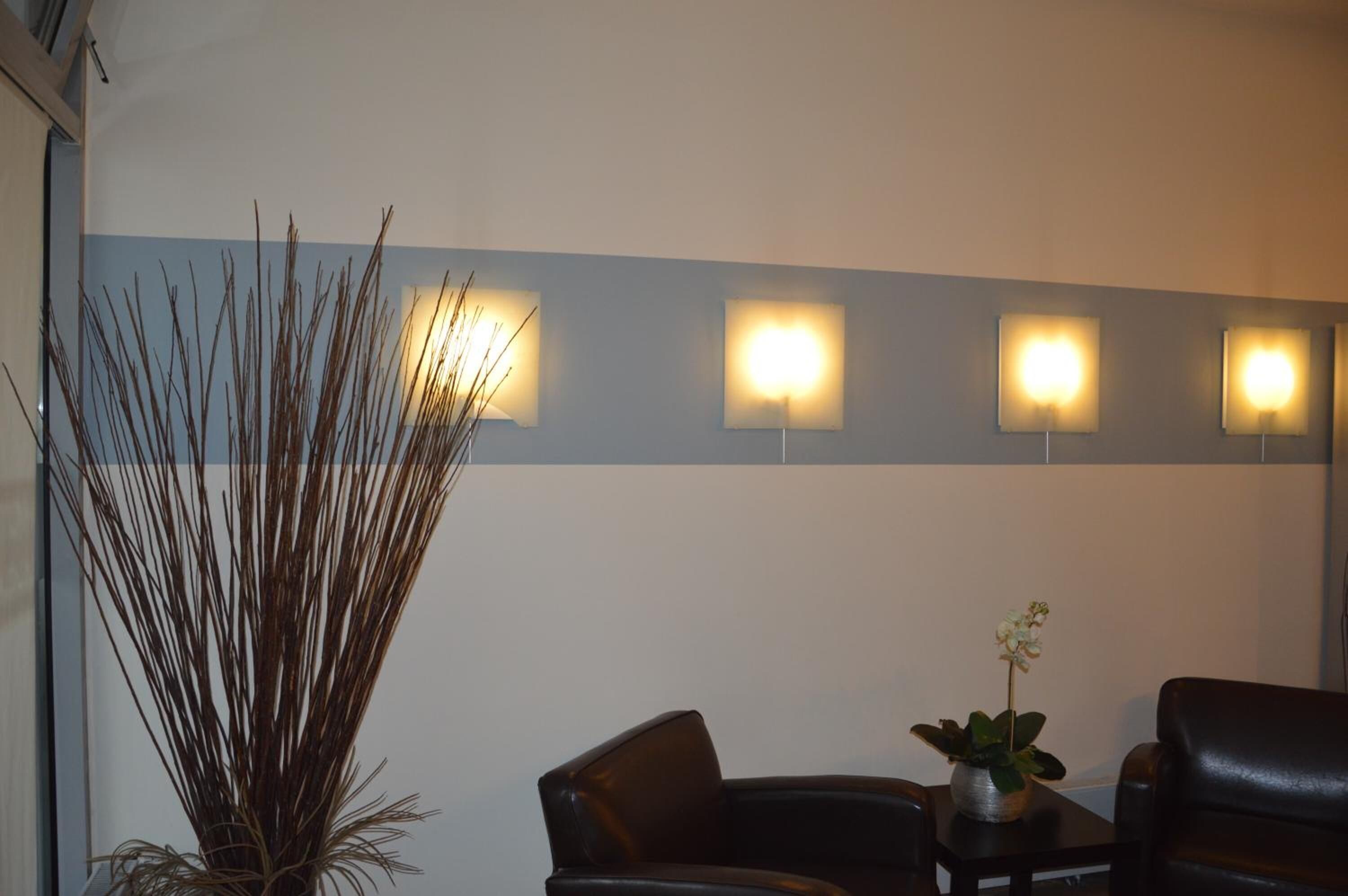 A waiting area with a tall plant, two leather chairs, a small table with a potted plant, and four wall-mounted lights.