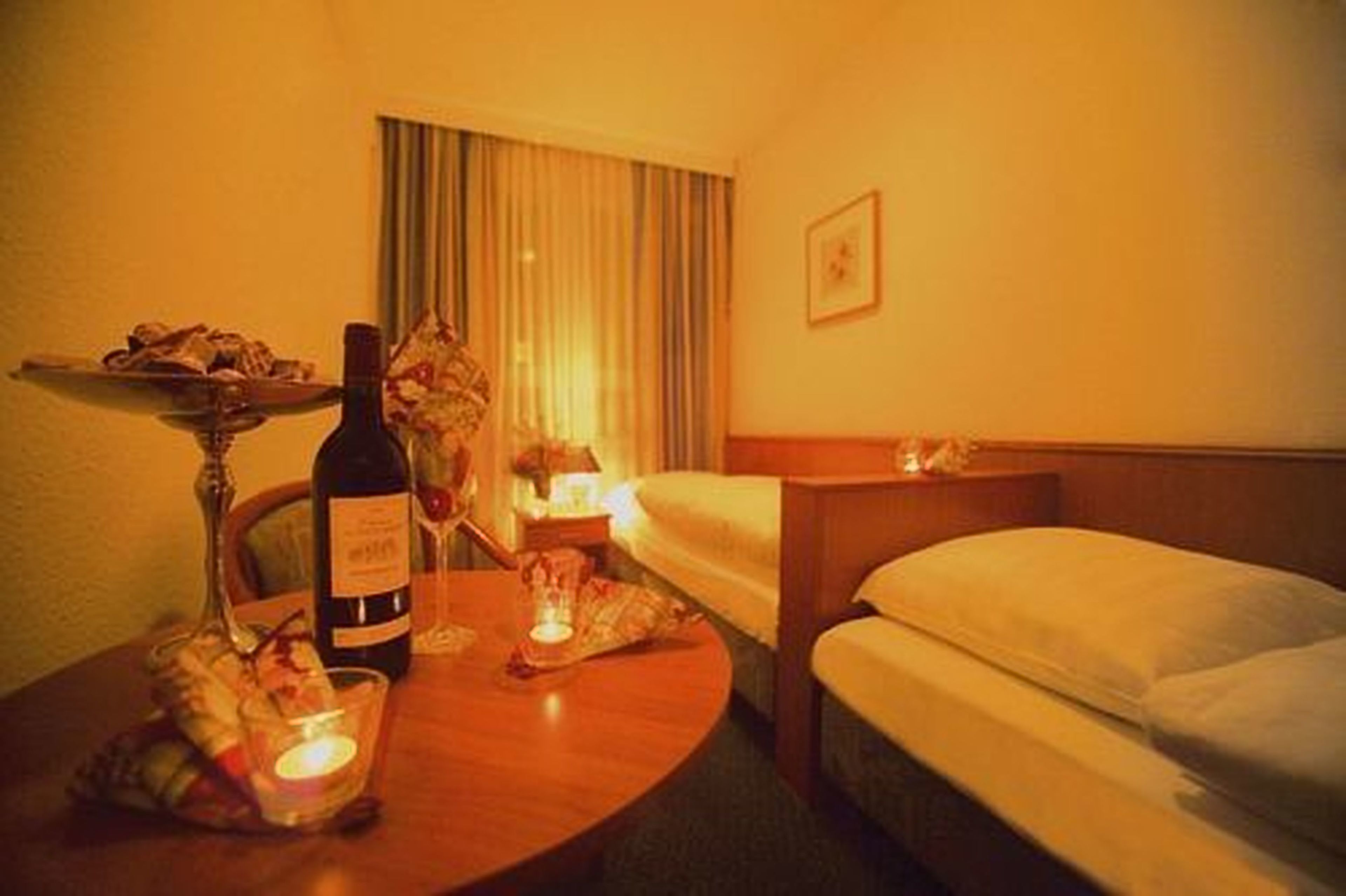 A hotel room with a bed, a bedside table with a bottle and glasses, a small table with a candle, and a window with curtains.