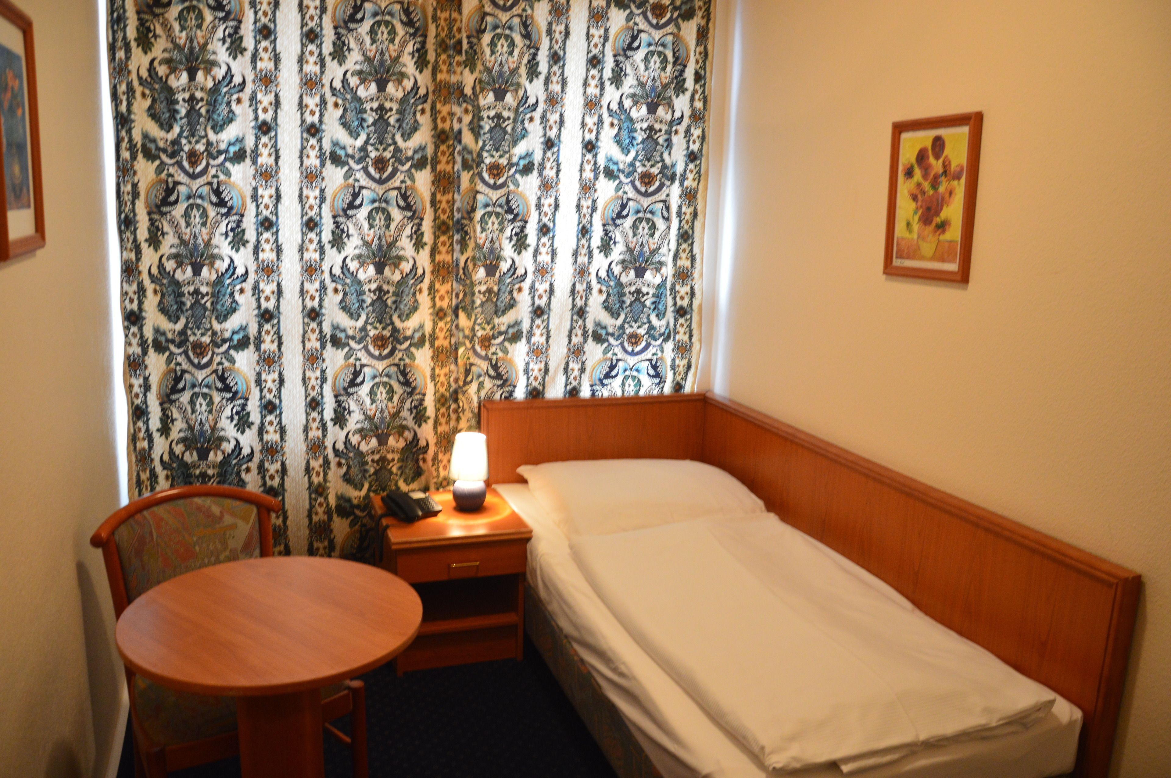 Une petite pièce, bien aménagée, avec un lit simple, une table de chevet avec une lampe, une chaise et un rideau orné d’un motif floral.