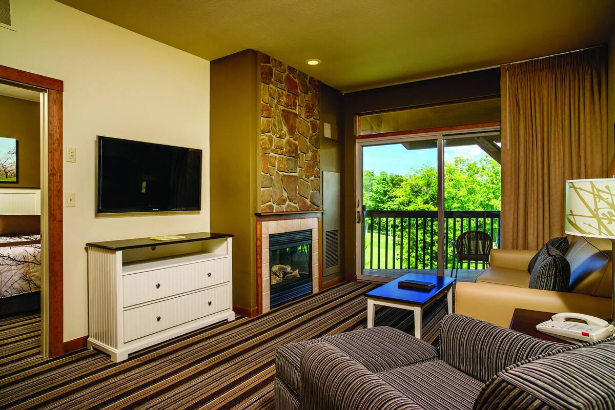 Une chambre d’hôtel dotée d’une cheminée en pierre, d’une télévision, d’un canapé et d’un balcon offrant une vue sur la verdure.