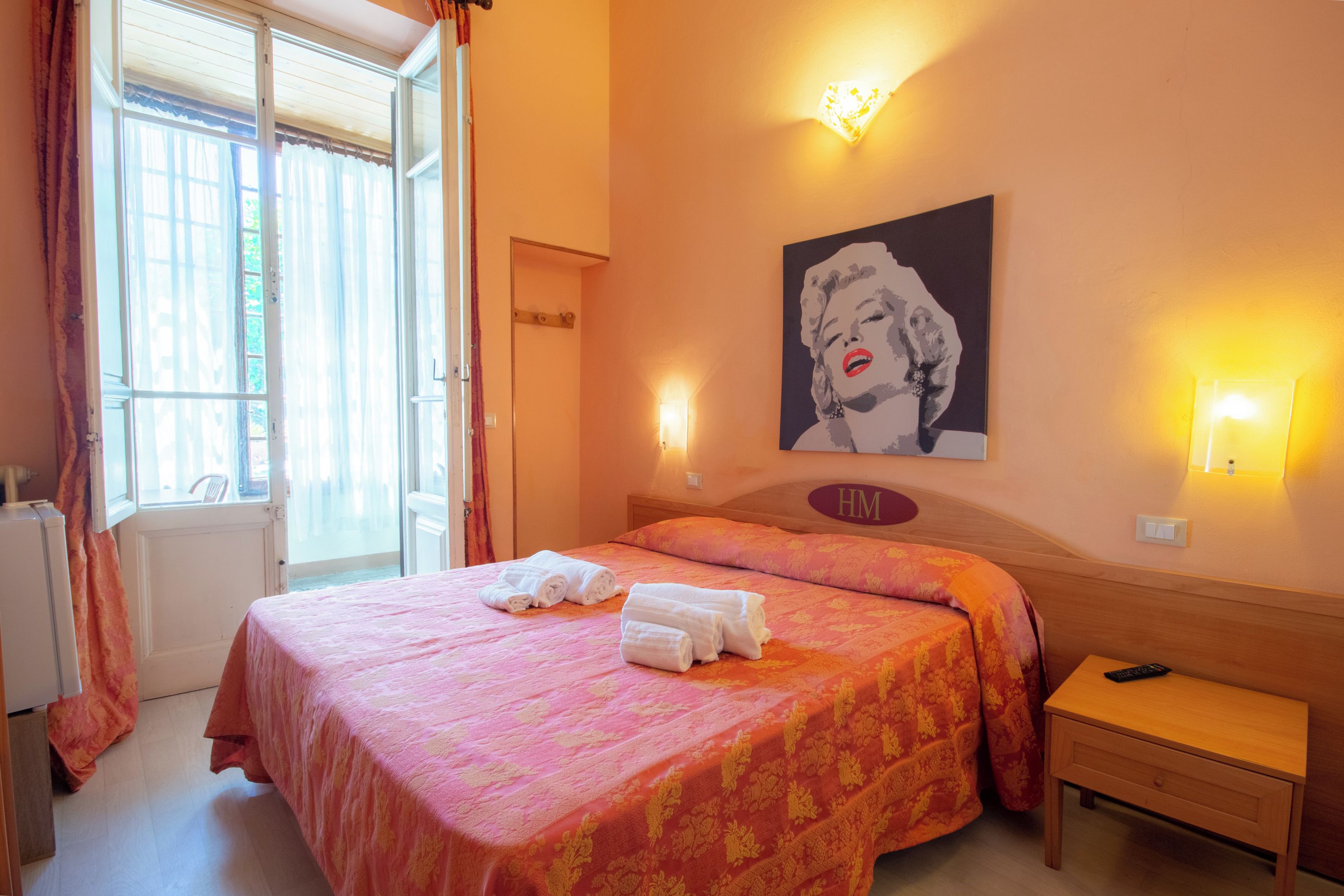A hotel room with a bed, a bedside table, a lamp, a painting on the wall, and a window with curtains.