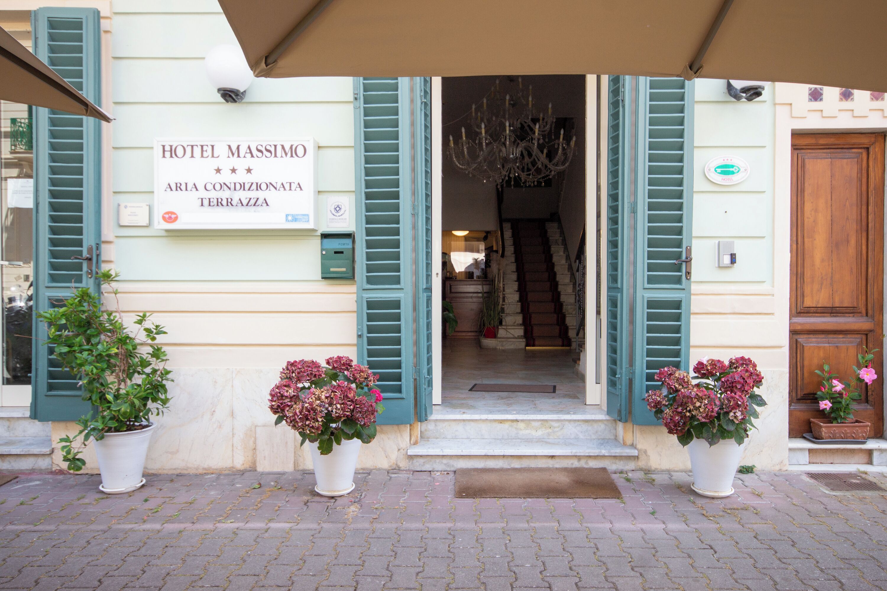 The entrance of Hotel Massimo with a signboard, a chandelier, and potted plants.