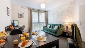 A hotel room with a dining table set with breakfast items, a green sofa, a lamp, and a suitcase. - Nemea Appart Hotel Toulouse Aéroport (Toulouse)