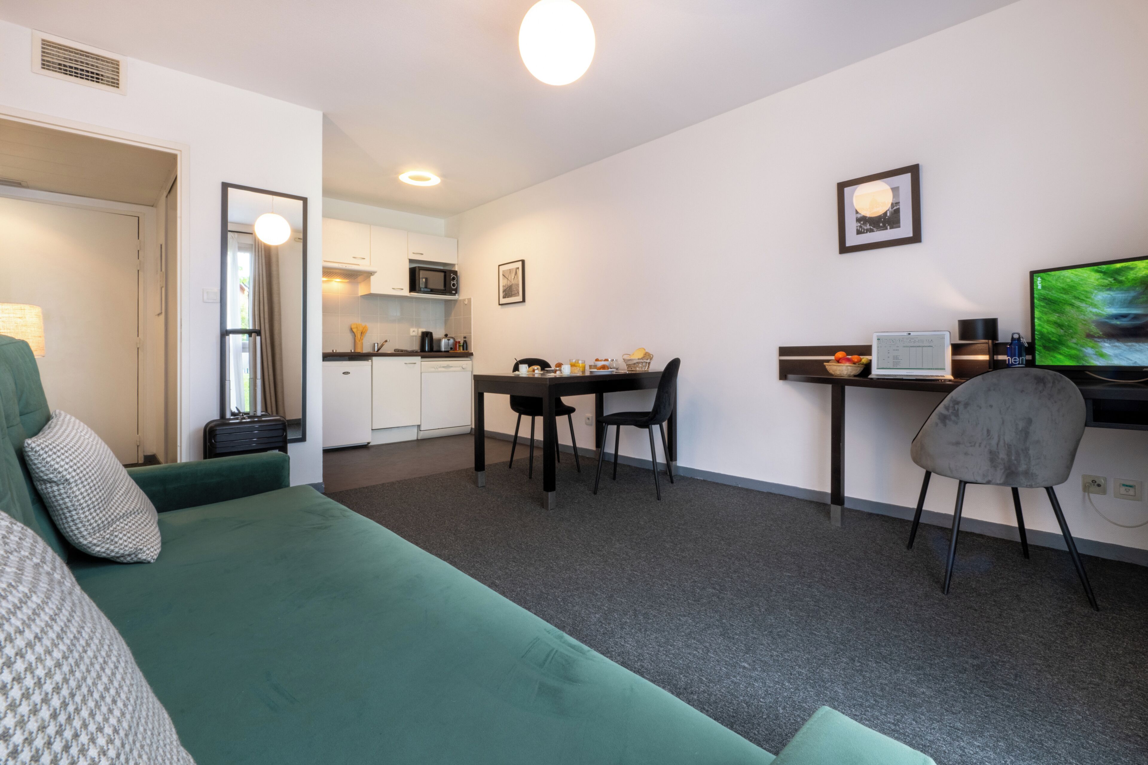 Un espace de vie compact comprenant un canapé vert, un coin repas avec une table et des chaises, un coin cuisine et un téléviseur posé sur un bureau.