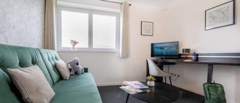 A room with a green sofa, a small table with a laptop, a chair, and a window with curtains.