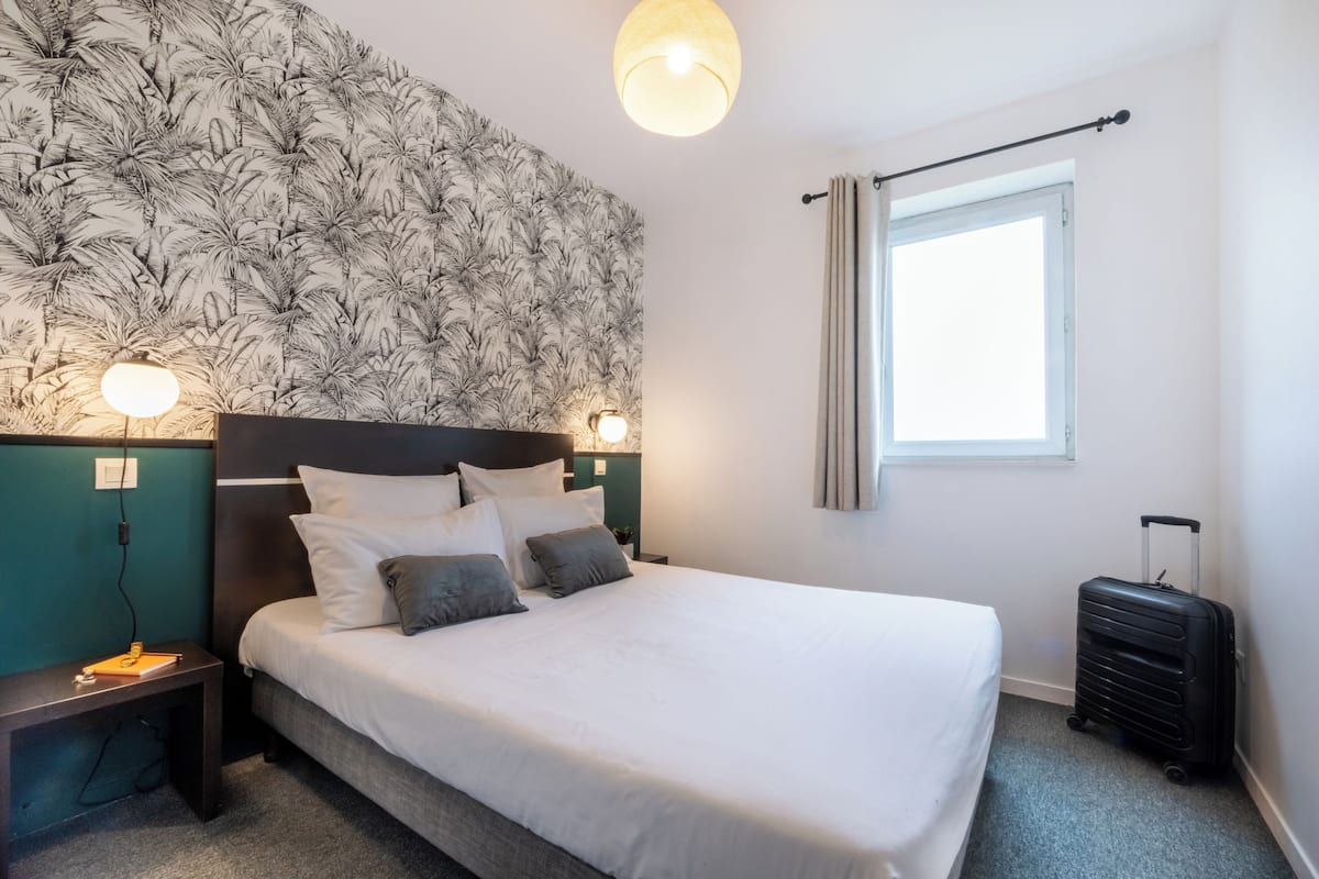 A neatly made bed with white and gray bedding, a headboard with a leaf pattern, two bedside lamps, a nightstand with a lamp, a window with curtains, and a suitcase.
