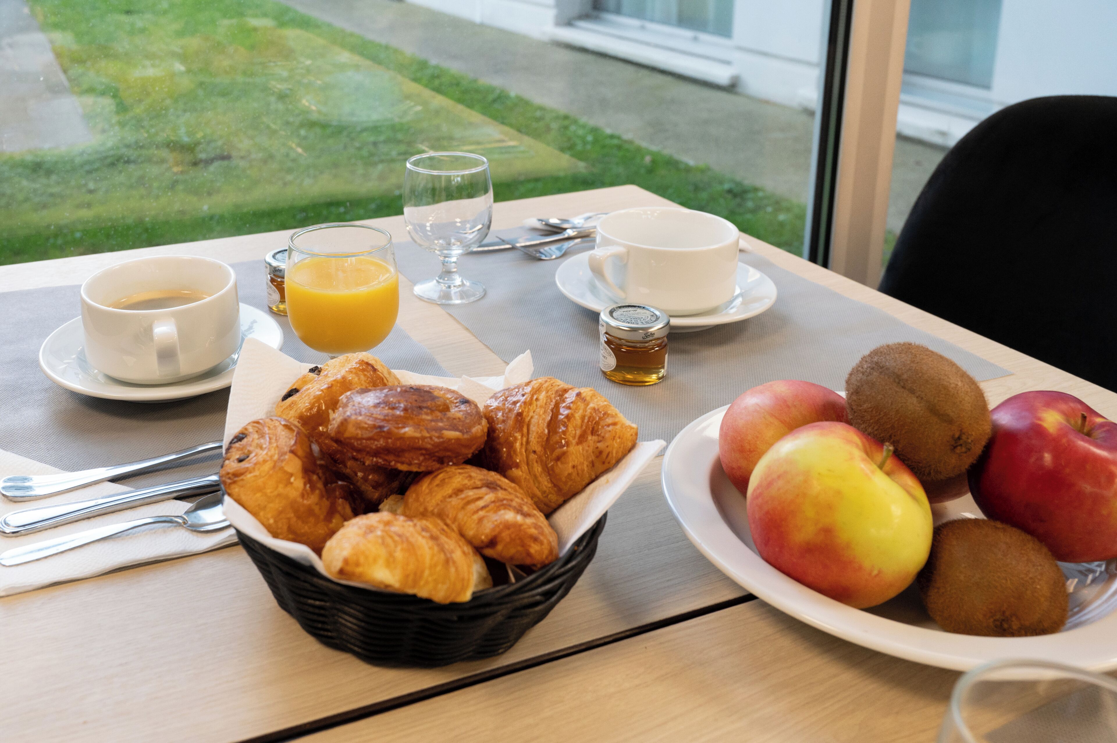 A breakfast table with croissants, apples, kiwi, orange juice, and coffee.