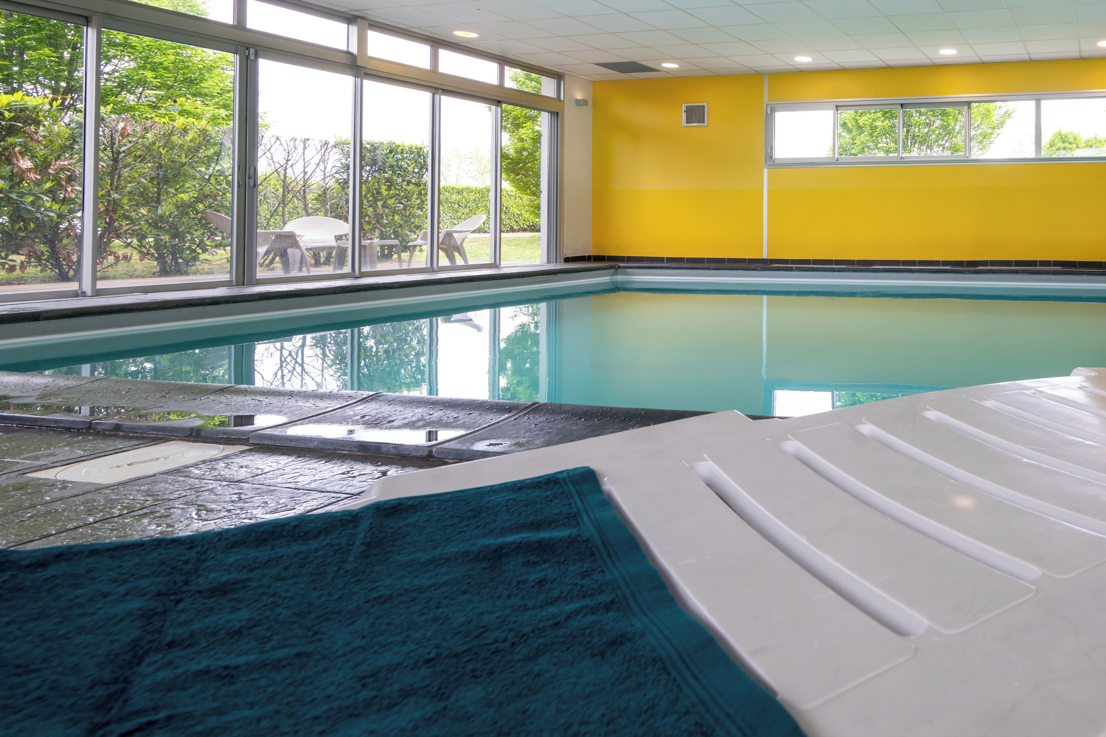 Piscina interior com paredes de vidro transparente, uma parede amarela e vista para o verde lá fora.