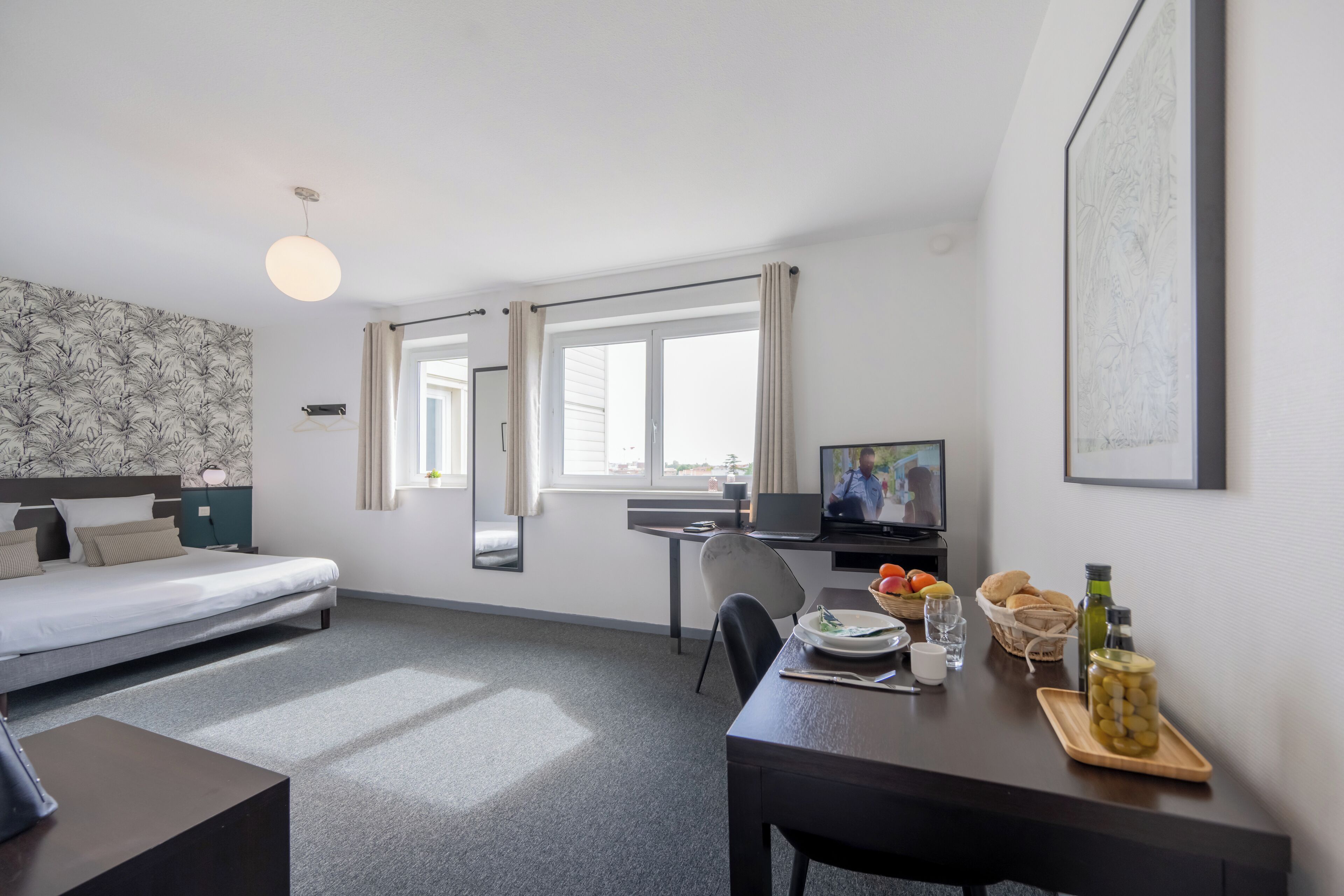 A hotel room with a bed, desk, TV, and breakfast table with food and drinks.