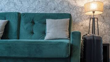 A teal sofa with two patterned pillows and a black suitcase against a floral wallpaper background.