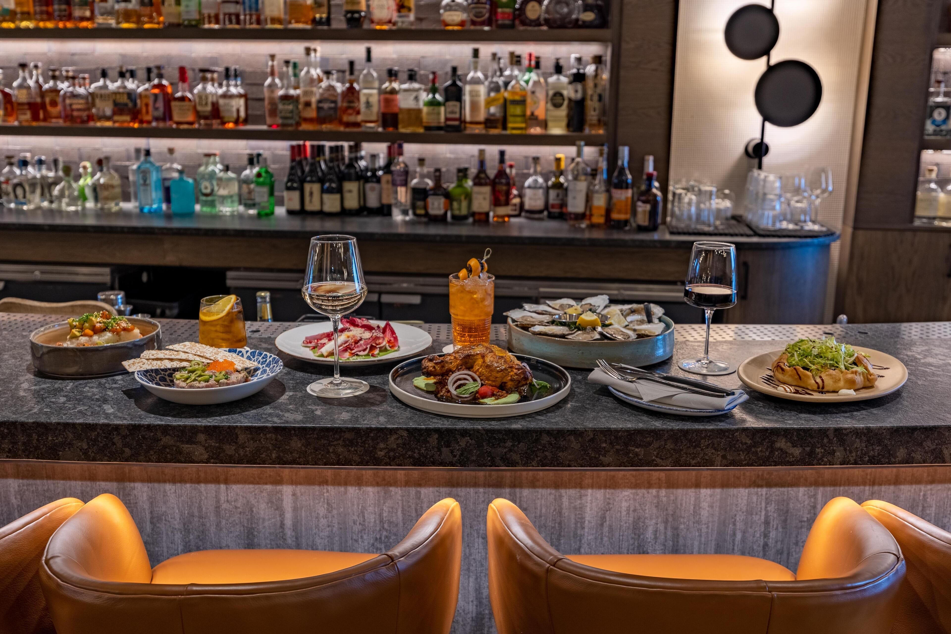 A bar area with various bottles and glasses, a counter with plates of food, and leather chairs.