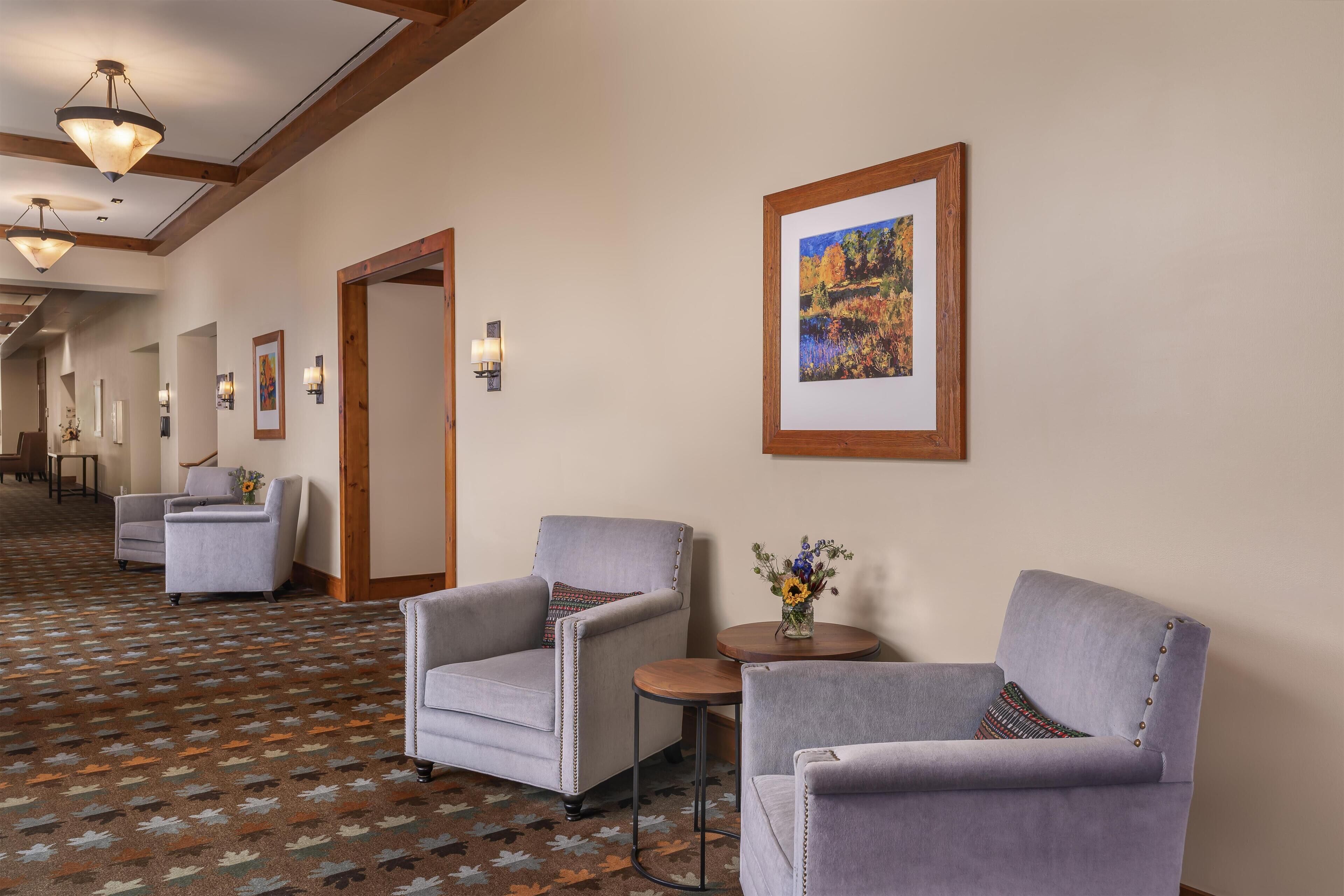 A hotel lobby with a patterned carpet, grey armchairs, a small round table with a vase, and framed artwork on the wall.