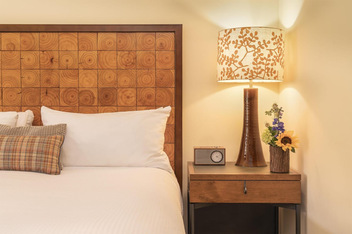 A neatly made bed with white and plaid pillows, a wooden headboard, a bedside table with a lamp, a radio, and a vase with flowers.