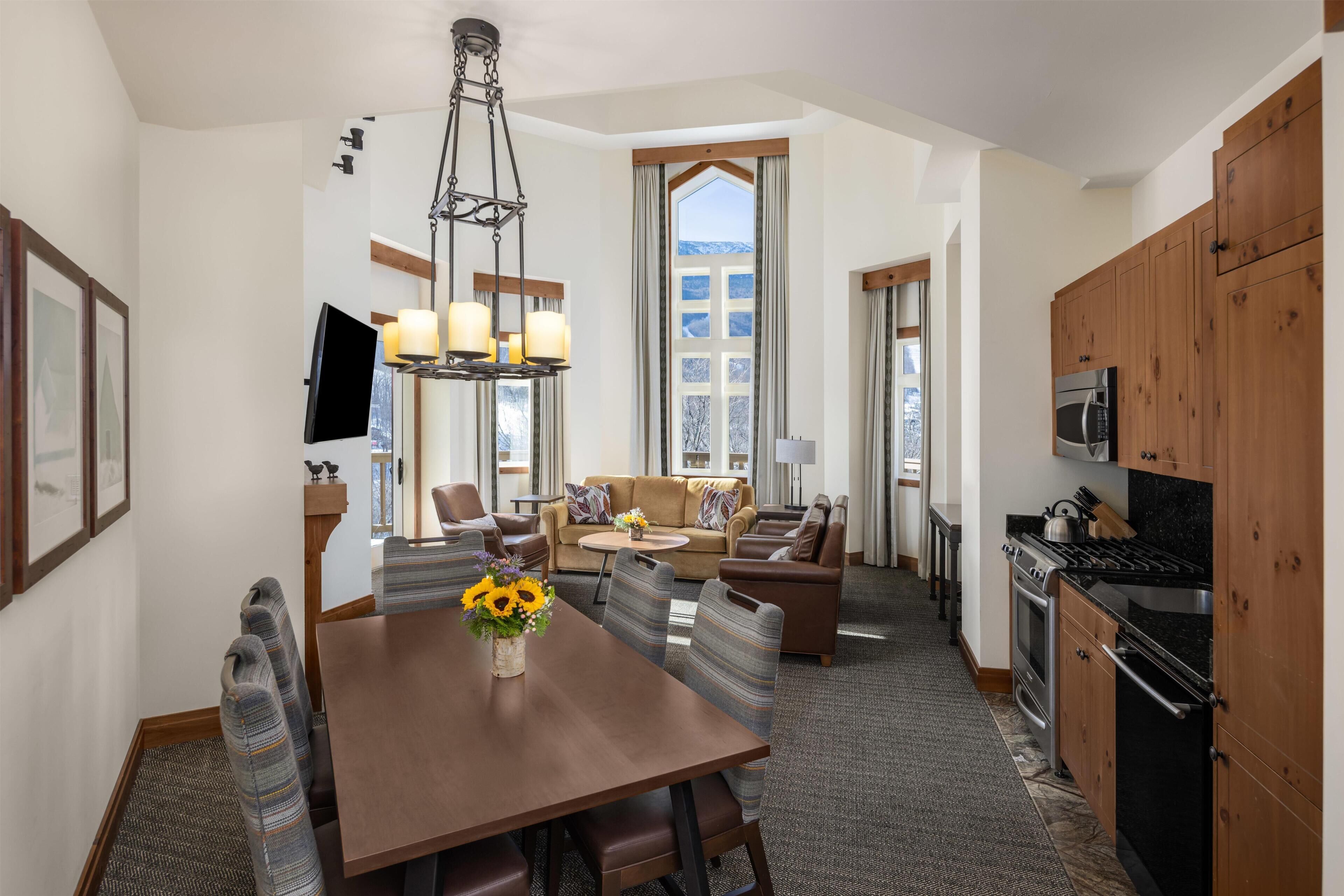 A modern hotel room with a dining area, a sofa set, a kitchen, and a large window with a view.