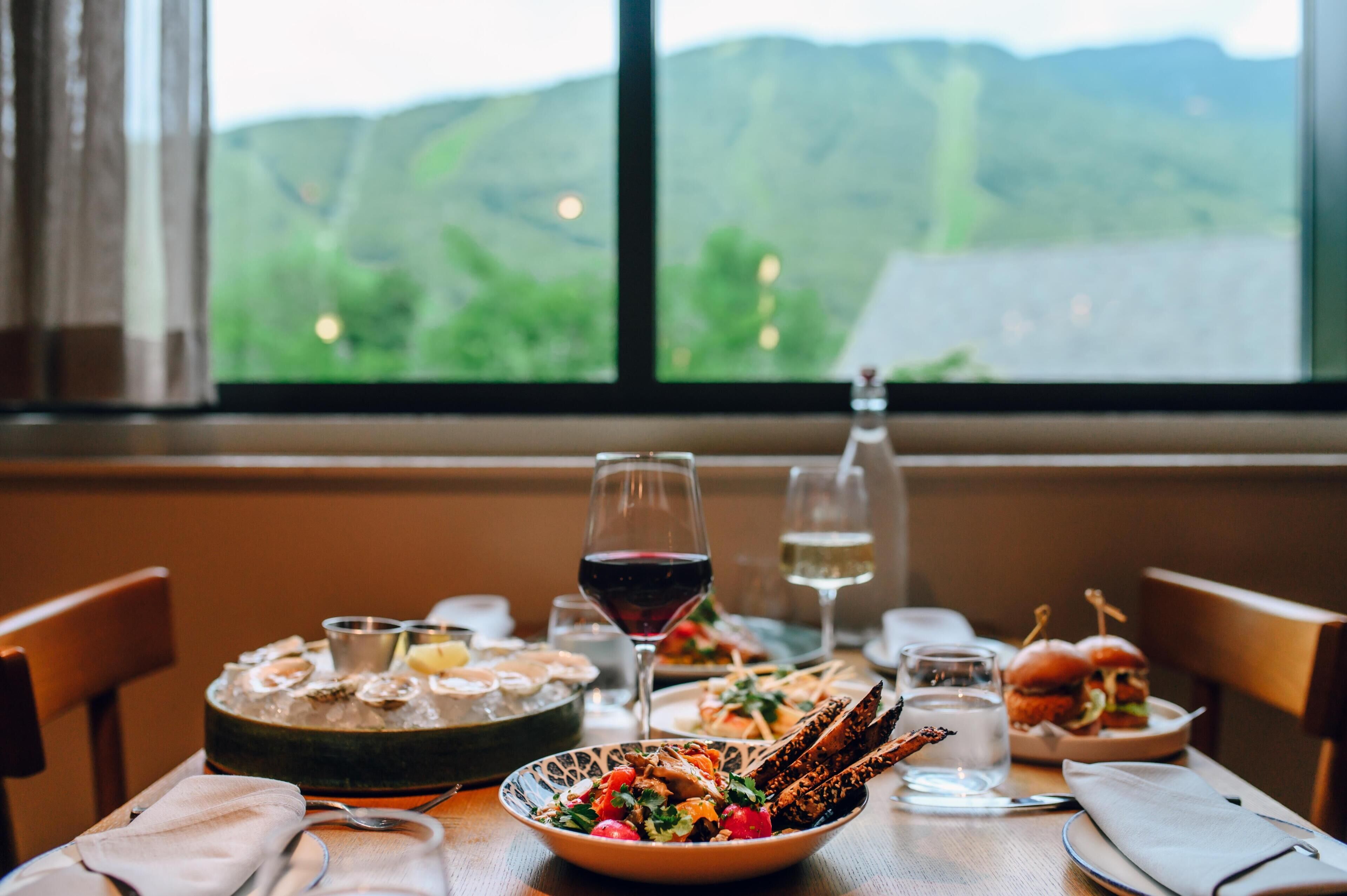Une table à manger dressée avec des fruits de mer, des hamburgers et des verres à vin, le tout mis en valeur par une vue panoramique sur les montagnes.