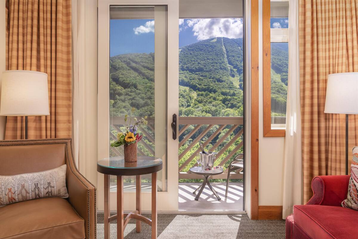 A room with a sofa, a small table with a vase of flowers, a view of a mountainous landscape through a door, and a balcony with a table and chairs.