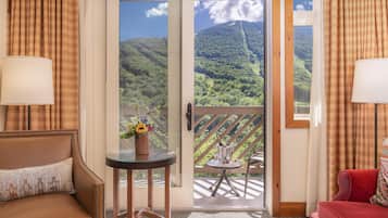 A room with a sofa, a small table with a vase of flowers, a view of a mountainous landscape through a door, and a balcony with a table and chairs.