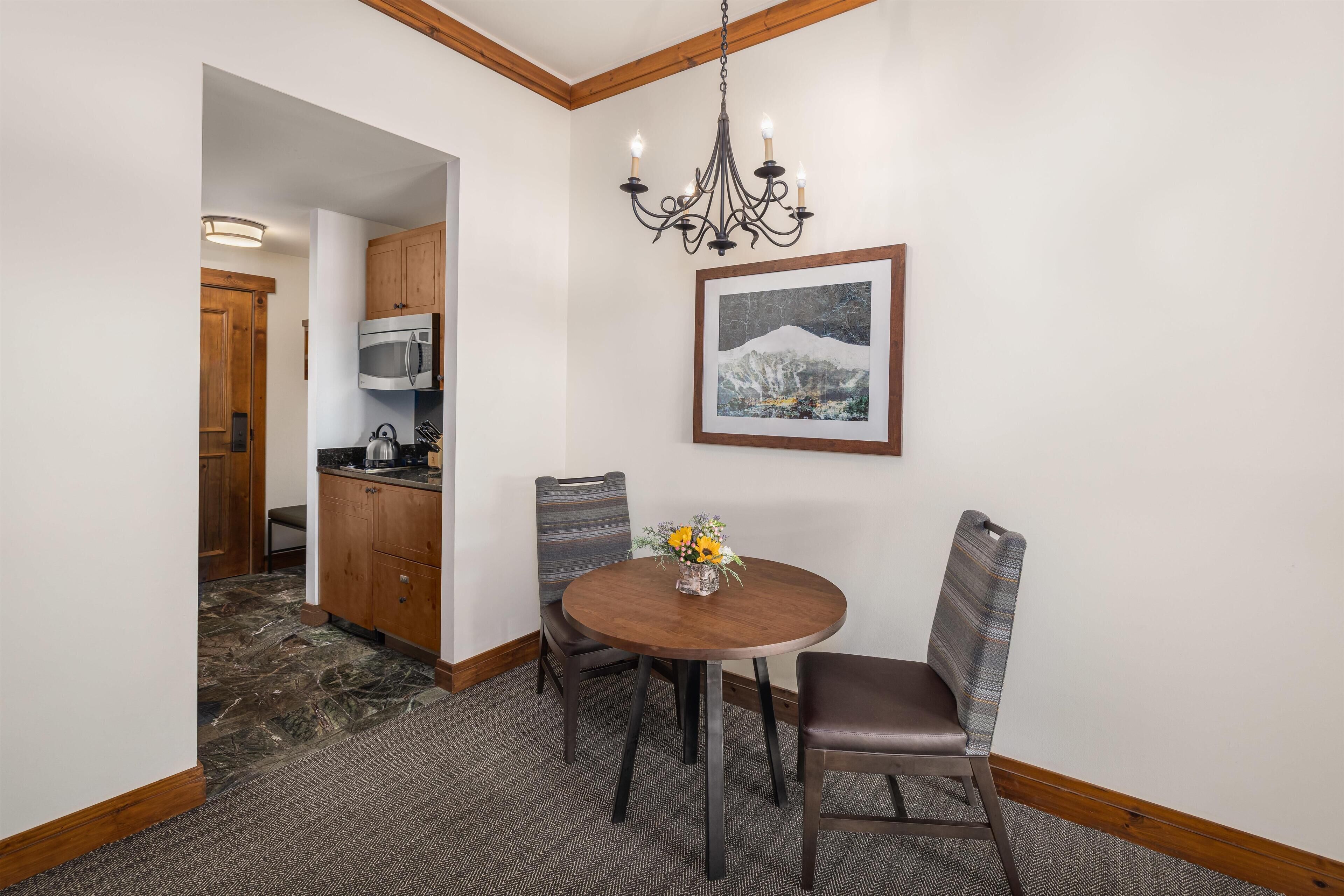Een eetgedeelte met een ronde tafel, twee stoelen, een kroonluchter en een lijst met een afbeelding aan de muur.