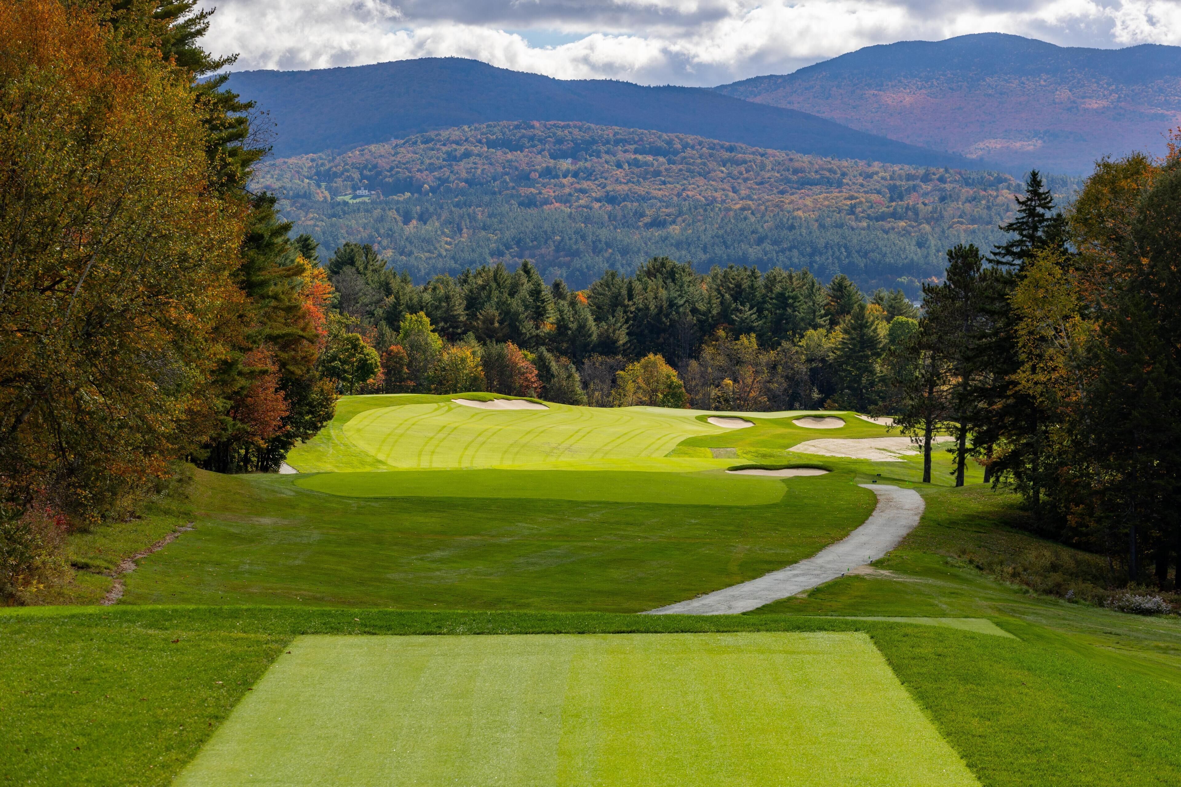 Un campo de golf con un sendero sinuoso, rodeado de árboles y montañas al fondo.