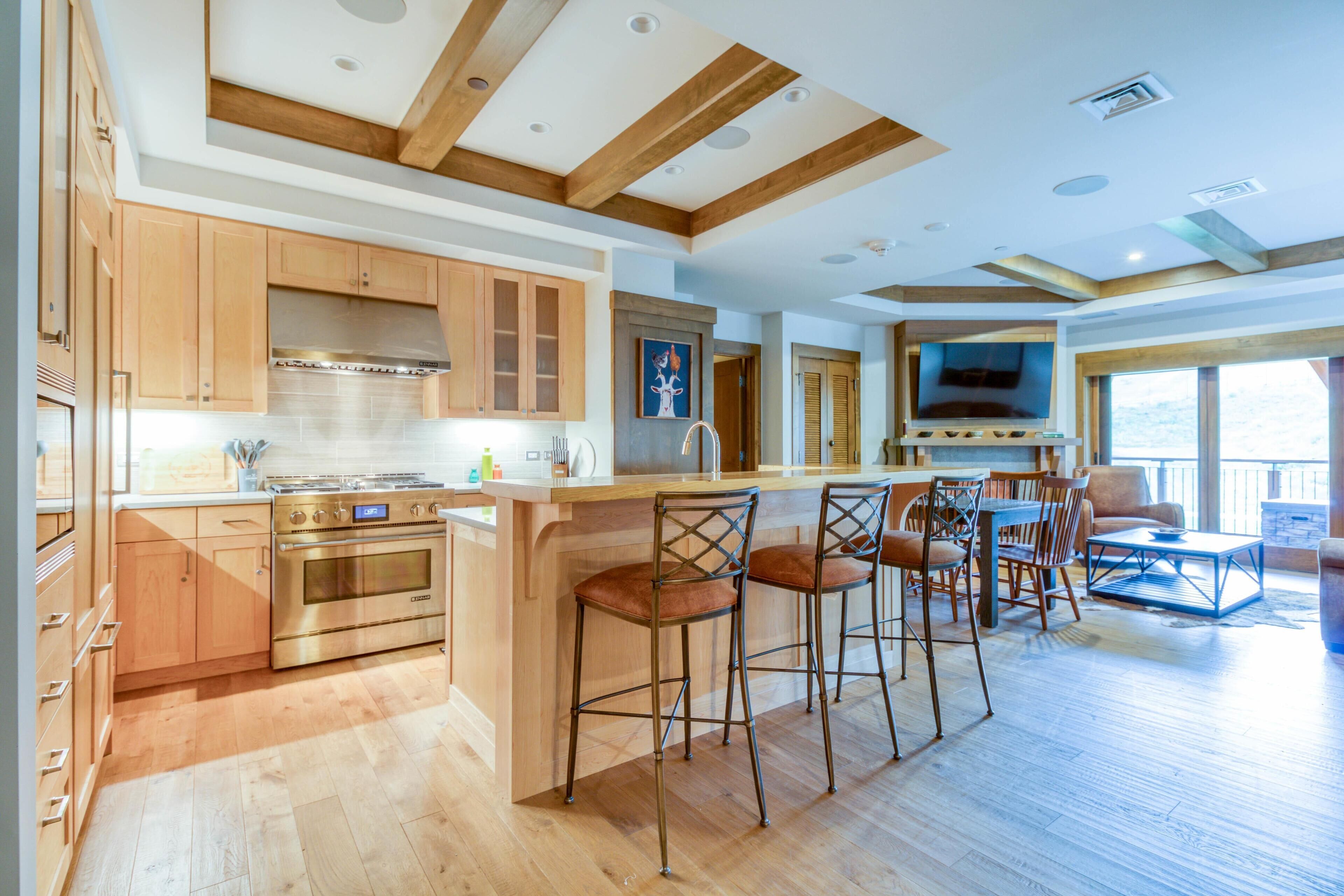 Een moderne keuken met houten kastjes, een centraal eiland met barstoelen, een eethoek met televisie en een groot raam.