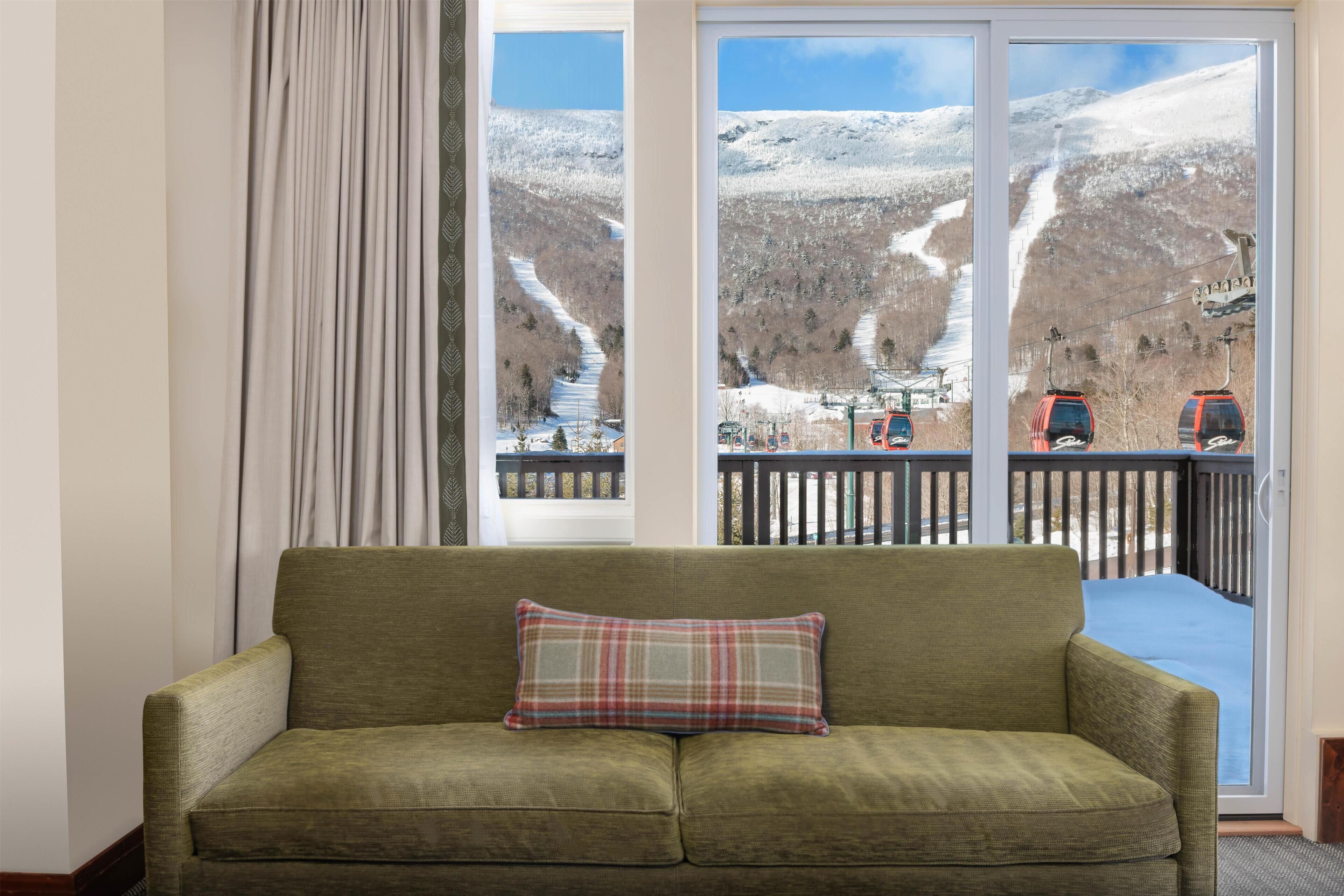 Een groene bank met een geruit kussen voor een raam met uitzicht op een besneeuwde berg en een skilift.