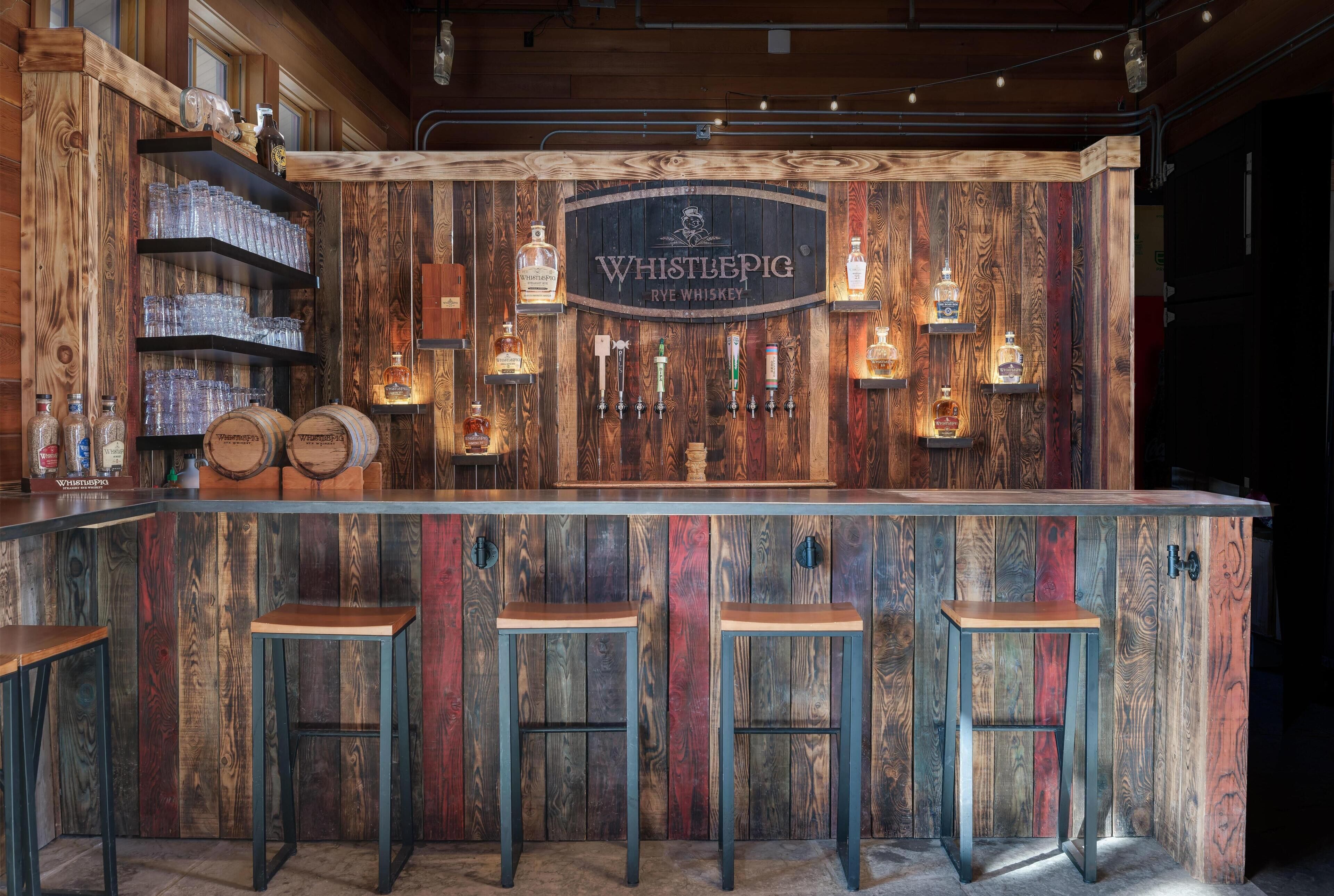 Un coin bar rustique avec un décor en bois, une enseigne indiquant « Whistlepig Rye Whiskey », ainsi que de nombreuses bouteilles et des verres.