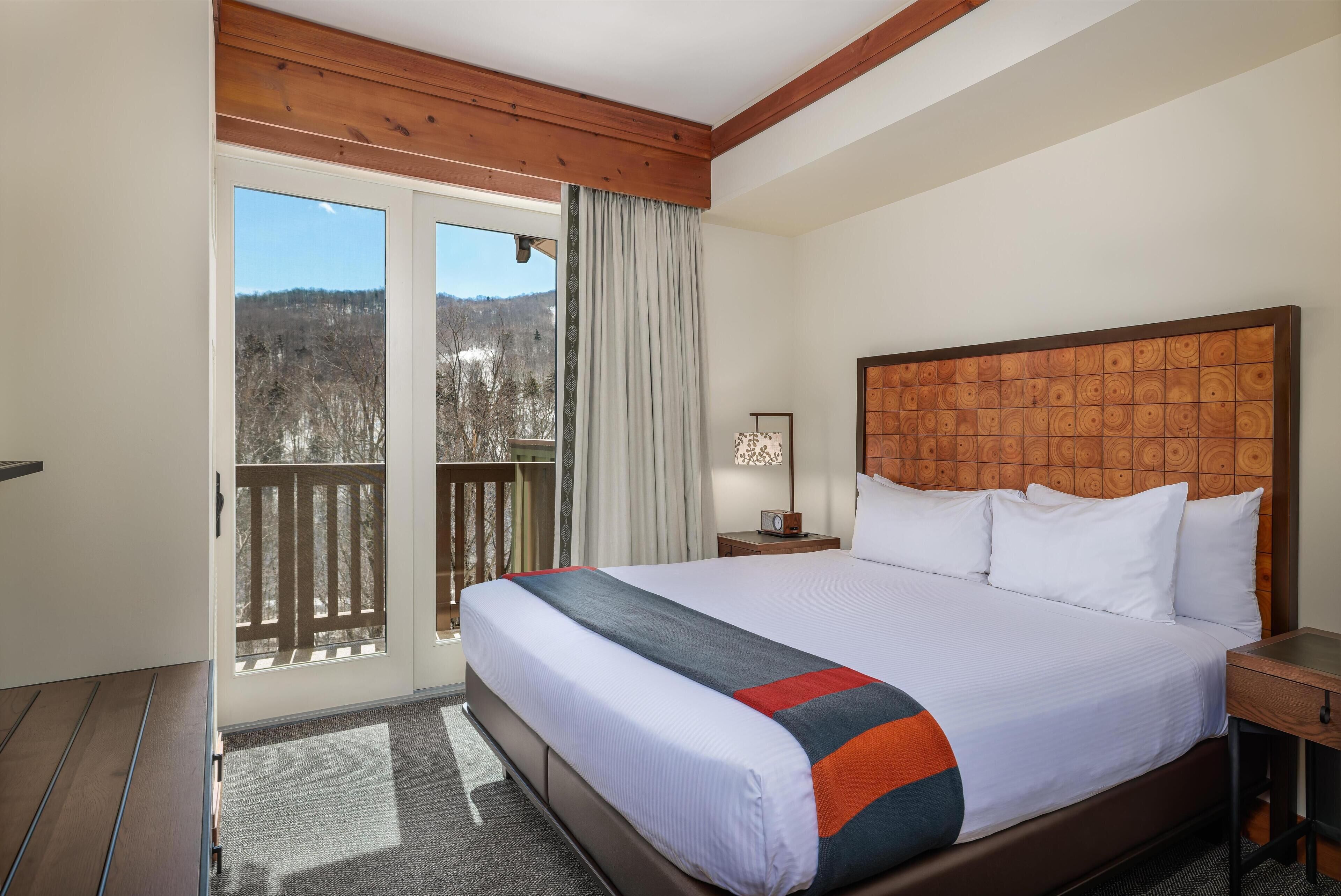 Une chambre d’hôtel avec un grand lit, une tête de lit en bois, une table de chevet, une lampe et une vue sur un balcon doté d’une balustrade en bois.