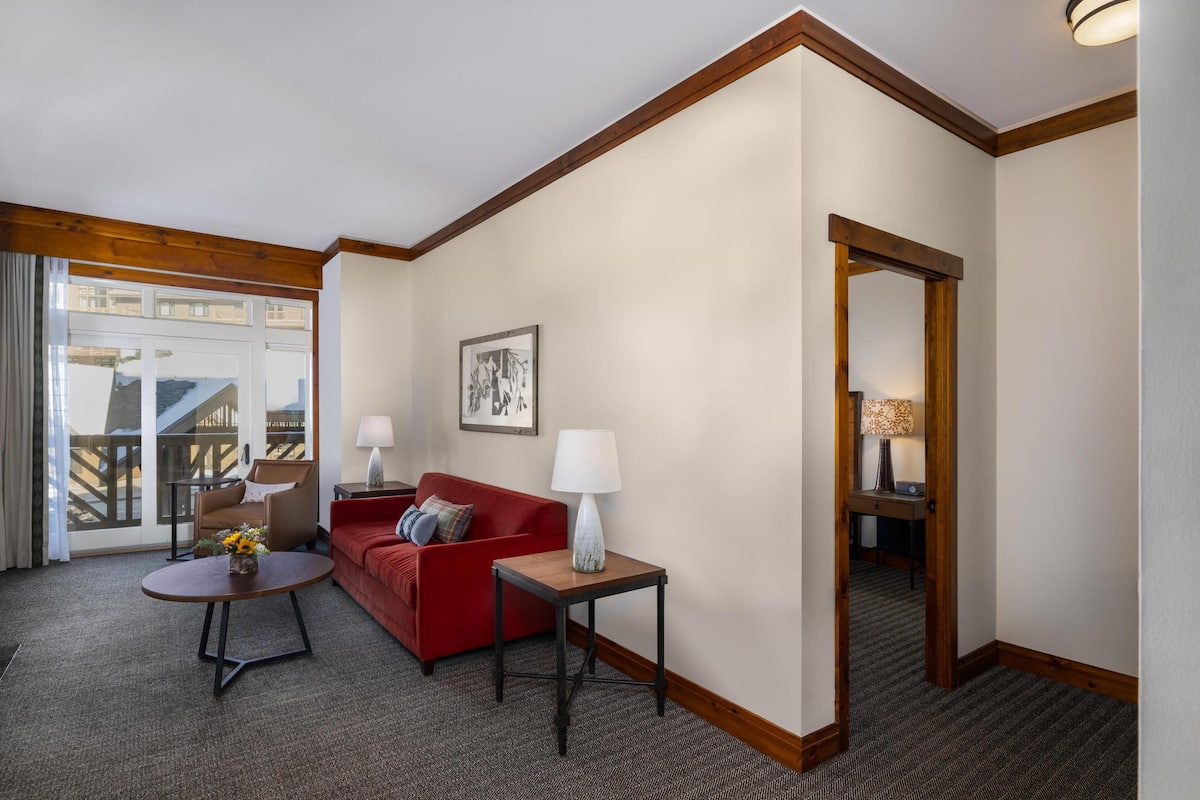 A hotel room with a red sofa, a small round table with a lamp, a framed picture on the wall, and a view through a glass door.