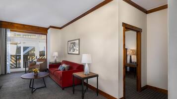 Quarto de hotel com um sofá vermelho, uma mesa redonda pequena com uma luminária, um quadro na parede e vista através de uma porta de vidro.