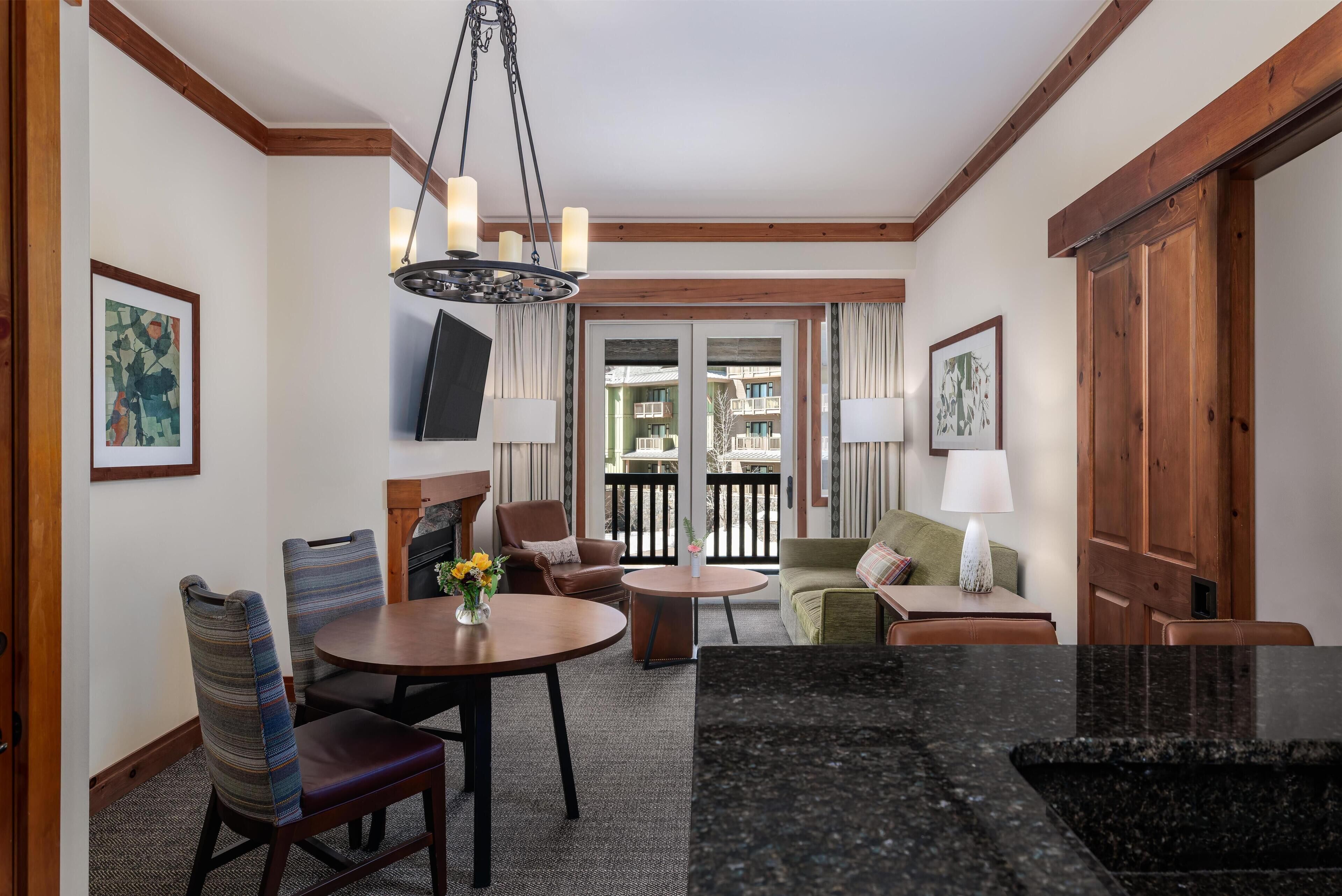 A dining area with a round table, chairs, a chandelier, and a view through a glass door to the outside.