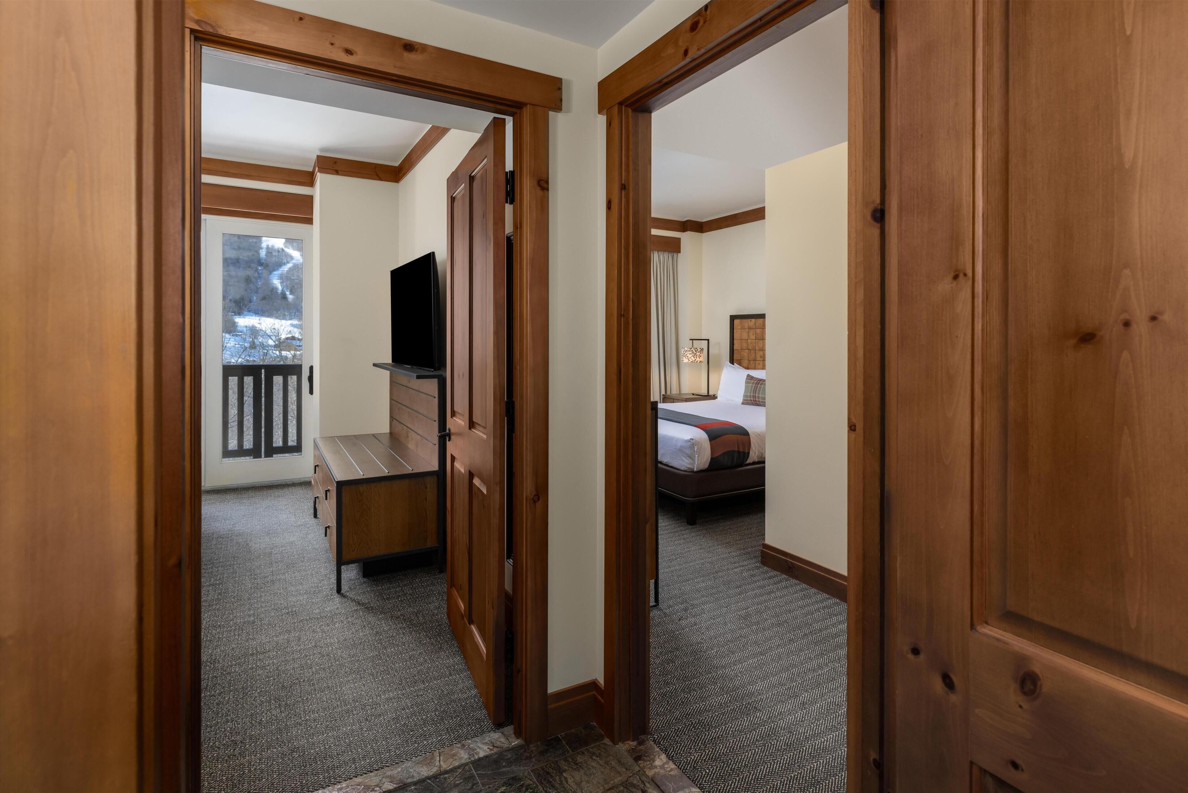 A hallway with wooden doors leading to a bedroom with a bed and a TV.