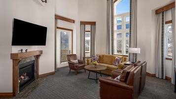 A hotel room with a fireplace, a flat-screen TV, a sofa, a coffee table, and large windows offering a view of the outdoors.
