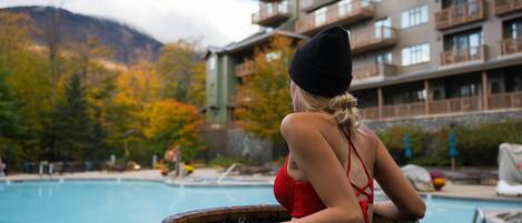 A woman in a red swimsuit and black beanie sitting on a wooden barrel by a pool with a mountain and lodge in the background.