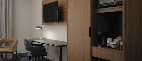 A hotel room with a desk, two chairs, a TV mounted on the wall, a microwave, and a coffee maker on a shelf.