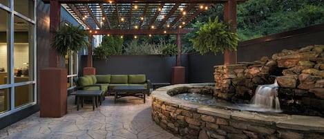 An outdoor patio with a stone fountain, a wooden pergola with string lights, and a green sofa.