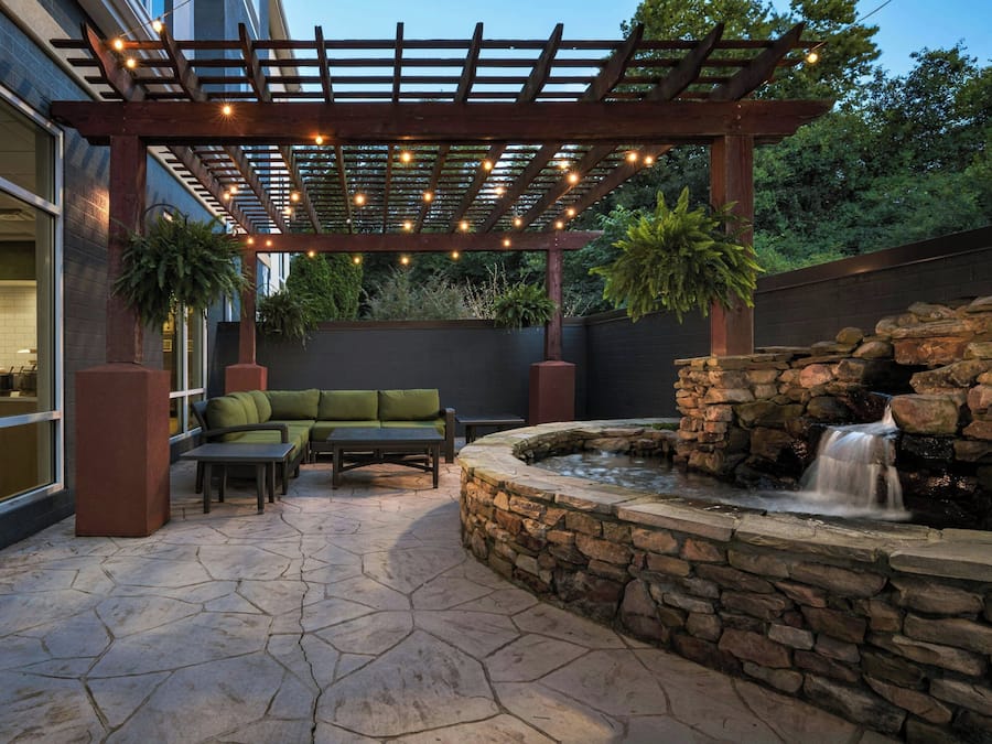 Une terrasse extérieure agrémentée d’une fontaine en pierre, d’une pergola en bois illuminée de guirlandes lumineuses et d’un canapé vert.