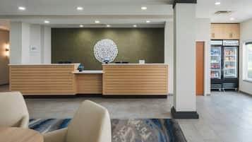 En hotellobby med modern design, med en receptionsdisk, ett dekorativt väggobjekt och ett minibar-kylskåp fyllt med drycker och snacks.