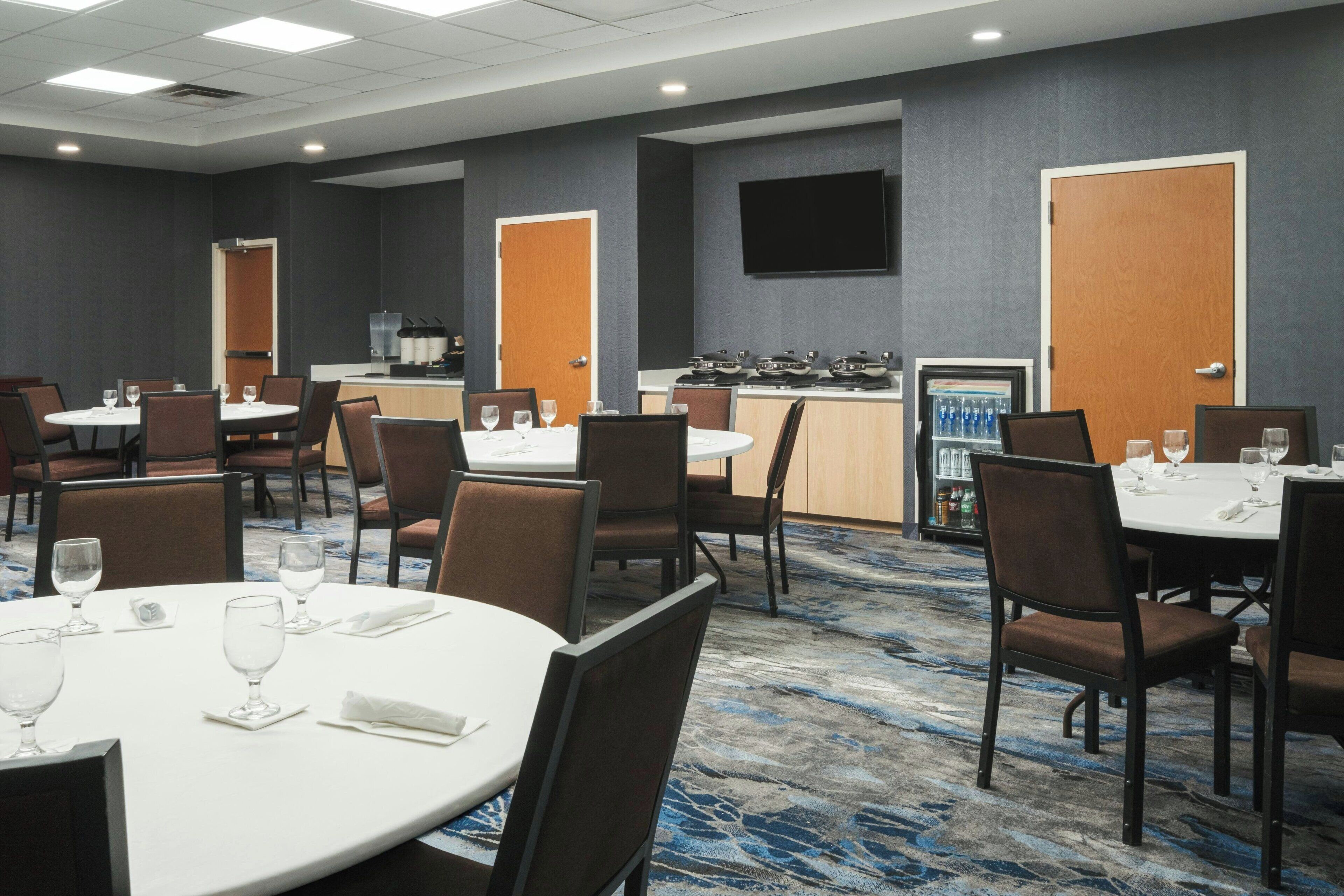 Comedor de hotel con mesas redondas, sillas y una zona de bar con refrigerador y bebidas.