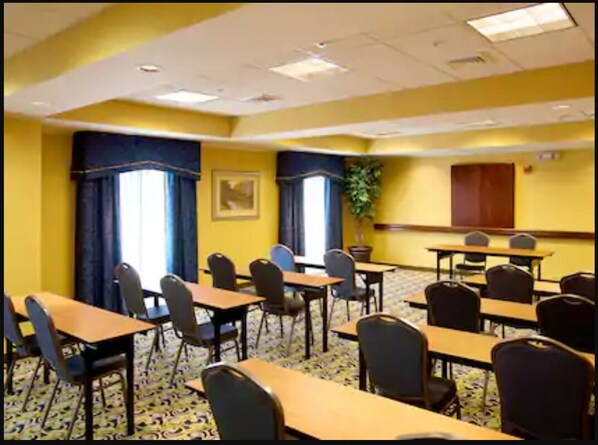 Un salón de conferencias con mesas y sillas, un ventanal con cortinas y una planta en la pared.