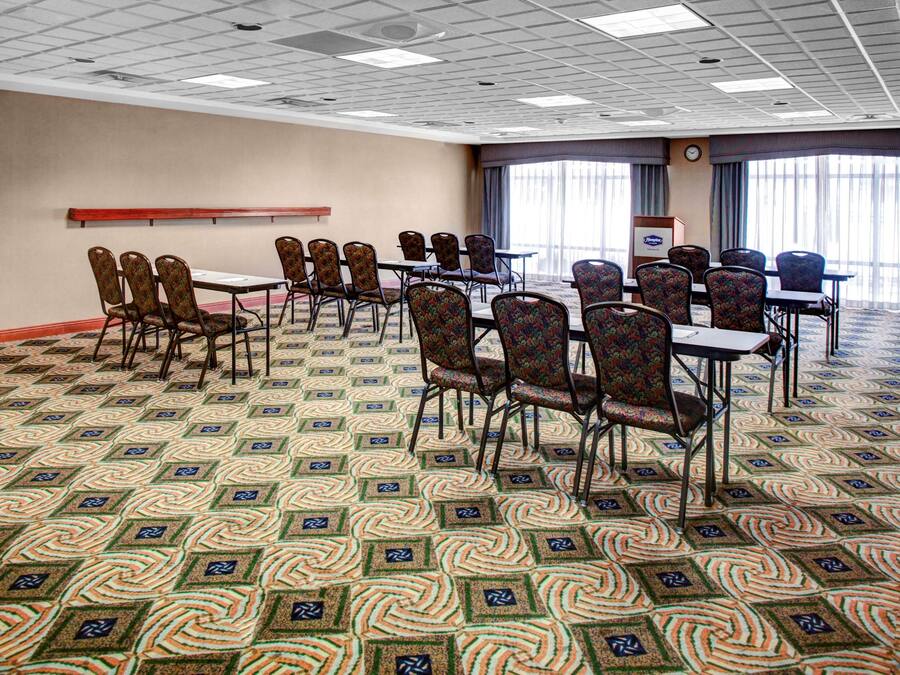 Un salón de conferencias con alfombra estampada, filas de sillas, mesas, un reloj y un ventanal con cortinas.