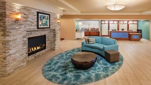 A living room with a stone fireplace, a blue sofa, a round coffee table, and a kitchen area in the background. - Homewood Suites by Hilton Virginia Beach/Norfolk Airport (Virginia Beach)