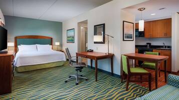 A hotel room with a bed, desk, chair, kitchenette, and a patterned carpet.