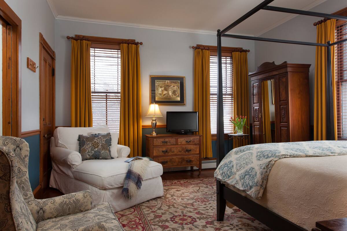 A bedroom with a four-poster bed, a sofa, a television on a wooden cabinet, and a floral-patterned rug.